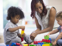 A teacher is sitting on the floor with her preschoolers and day care students - they are playing with plastic blocks together.